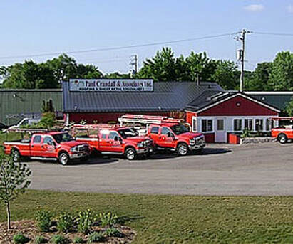 A picture of the Paul Crandall & Associates store front during the summer. The picture features the red shop along with three signature red trucks parked at an angle.