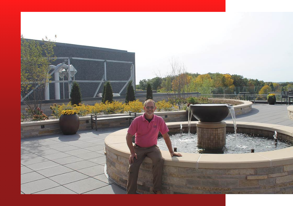 Paul Crandall of Paul Crandall and Associates, Inc. sitting on a fountain, which is a part of the custom green roof system installed for ProHealth Care. The picture features yellow flowers, green trees, benches, planters, and the fountain.